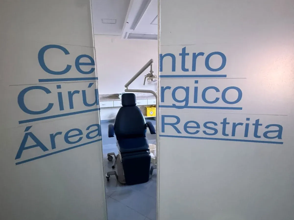 Porta de vidro com a inscrição “Centro Cirúrgico Área Restrita” e, ao fundo, a cadeira de procedimentos.